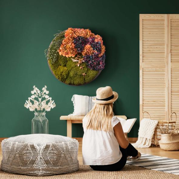 Rundes Moosbild aus Hortensien, Pflanzen und Waldmoos in einem Wohnzimmer und im Vordergrund eine lesene Frau 