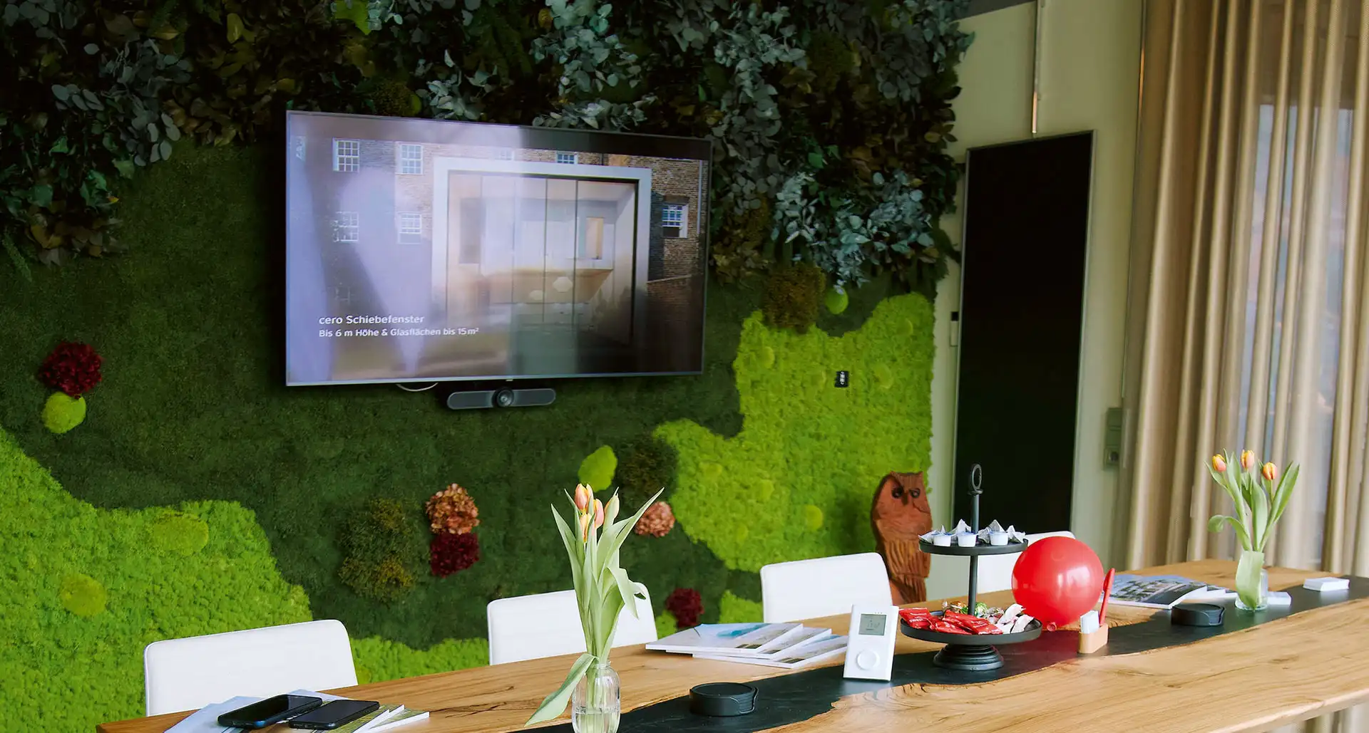 Heller Konferenzraum mit großflächiger Mooswand, Fernseher und Holztisch, kombiniert mit frischen Blumen und dekorativen Elementen für eine natürliche Arbeitsatmosphäre.