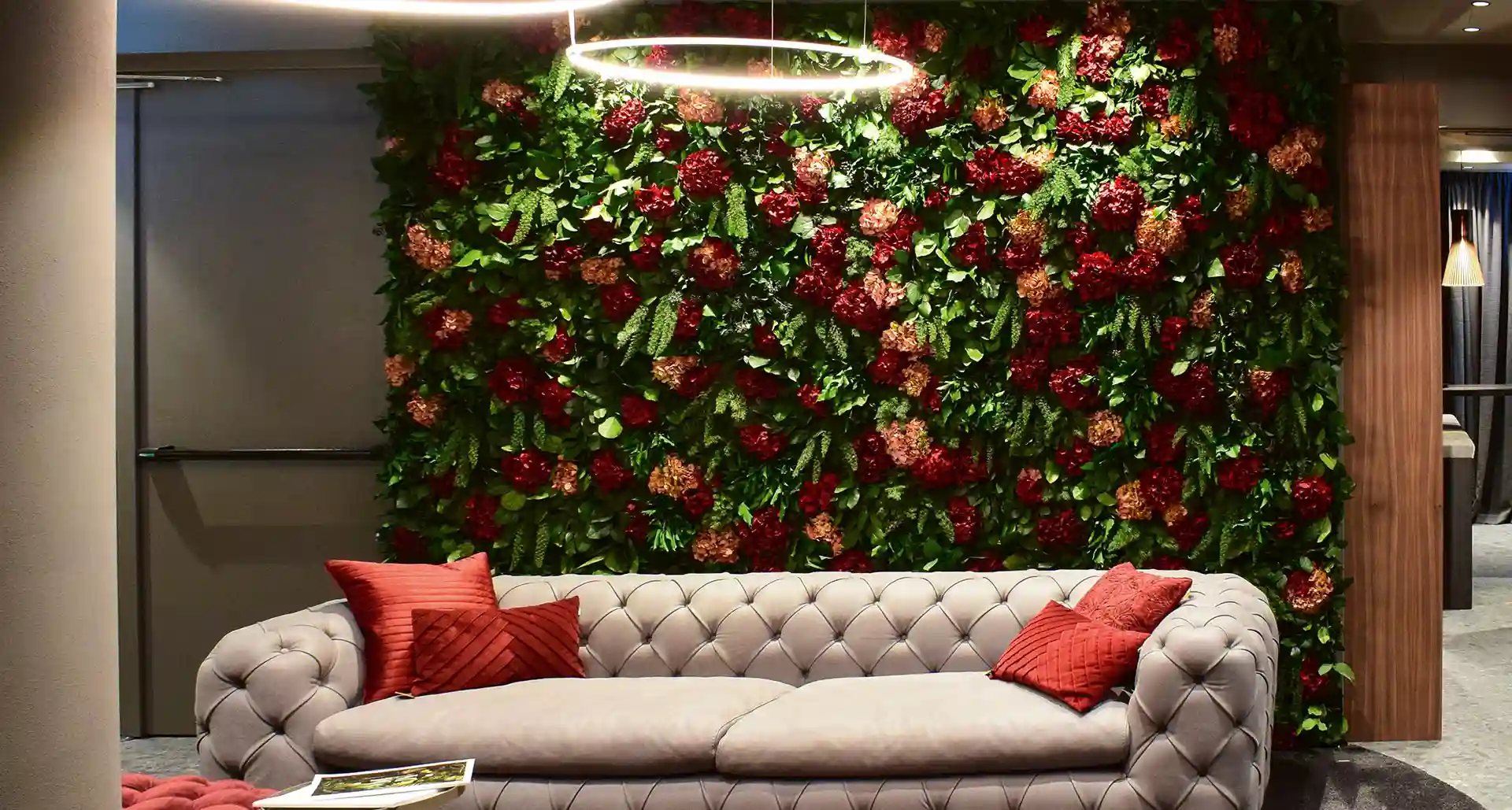 Stilvolle Lounge mit großflächiger Blumenwand aus roten und rosafarbenen Blüten, kombiniert mit einem hellen Sofa und roten Kissen für ein natürliches und elegantes Ambiente.